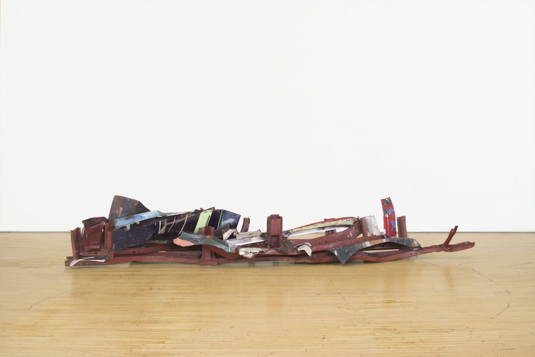 A long, low-lying sculpture made of red, dark blue, and green metal parts rests on a wooden floor.