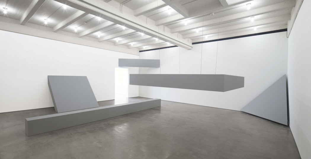 Five gray sculptures are installed in a gallery. From left to right, they include a long beam placed on the floor in front of a rectangular slab resting against a wall, a beam suspended in the left corner above a door, another rectangular slab suspended from the ceiling, and an equilateral triangle placed in the right corner.