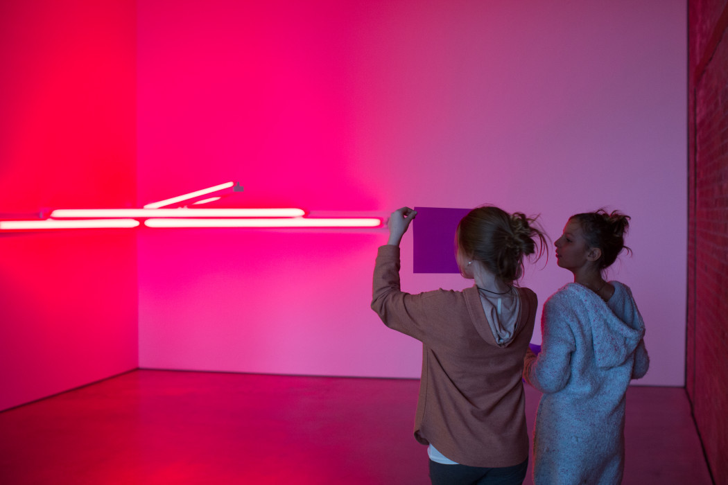 Two visitors look at a piece of paper that one holds in front of her face within a room made fuchsia pink by the light installation situated in the corner.