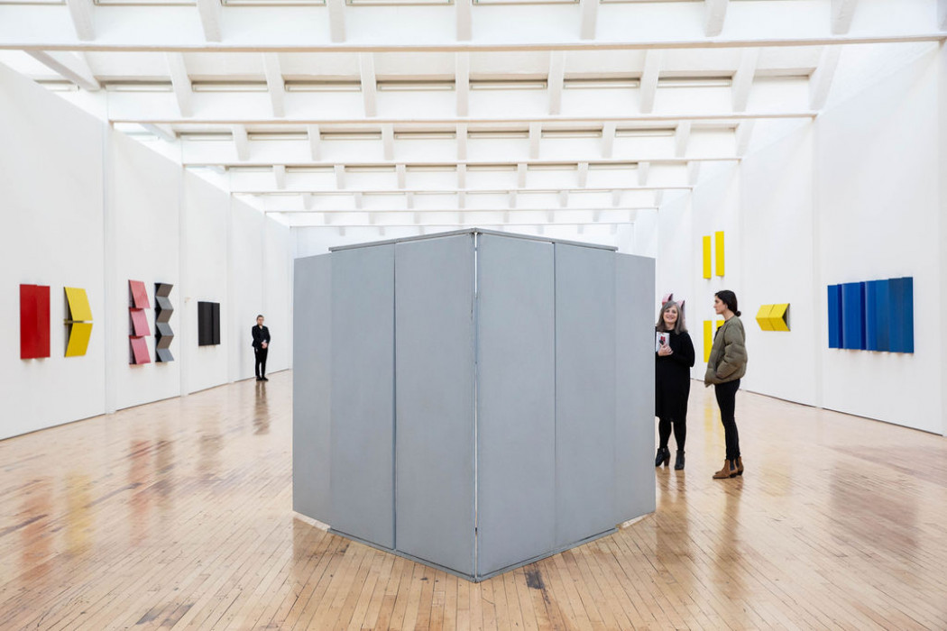 In the middle of a gallery displaying bright monochromatic hanging sculptures stands a grey box constructed from vertical panels about the size of the two visitors onlooking.