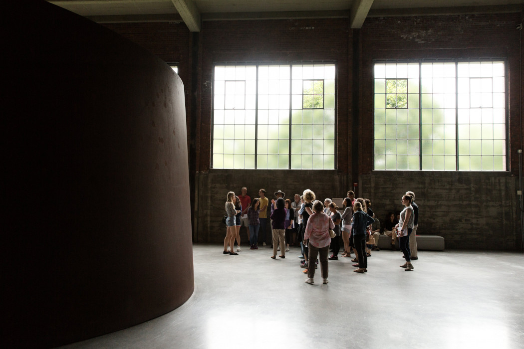A group of visitors gather in large space lit by two large windows behind them. A tall, curved steel sculpture is to the left.