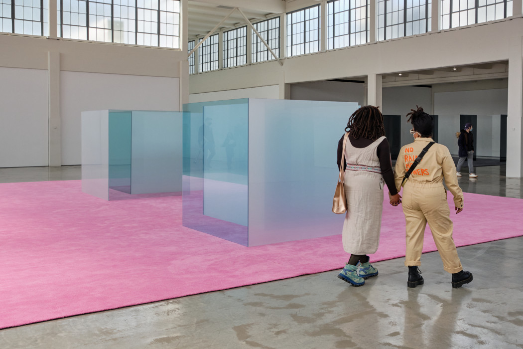 Two people hold hands as they walk along a bright pink carpet. Reflective blue and opaque glass box-shape sculptures sit in the middle of the carpet.