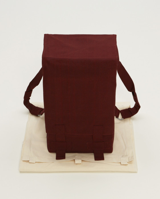 A rectangular object with two straps toward the back is wrapped in red fabric and placed above a folded beige muslin. 