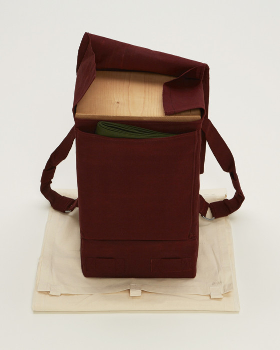 A rectangular object with two straps is wrapped in red fabric and placed above beige muslin. The objectÕs top flap is open, revealing a wooden slab and folded green cloth.