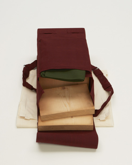 A rectangular object with two straps is wrapped in red fabric and placed above beige muslin. The objectÕs top flap is open, revealing three wooden slabs and a green cloth.