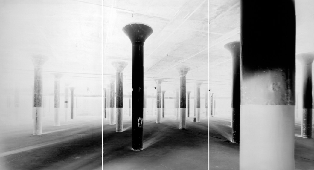 An inverted black and white photo of evenly spaced columns with flared ceiling tops in a large space.