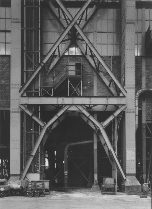 Black-and-white photograph of a glass and brick factory building behind a large metallic scaffolding.