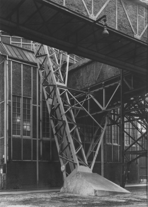 Black-and-white photograph of brick factory building featuring an open-air diagonal metal support structure and an aboveground upper-story brick passageway.
