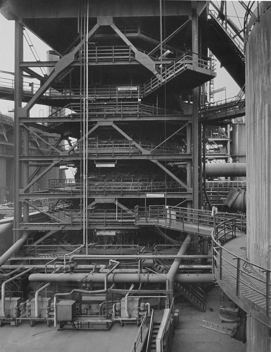 Black-and-white photograph of outdoor tiered metal structure with multilevel railings and walkways and large cylindrical pipes connecting to other structures.