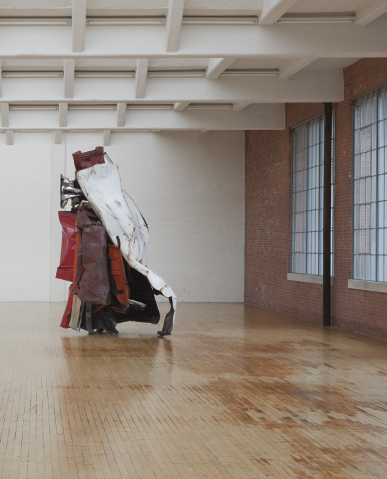 An oblong red, white, and silver sculpture made of metal is placed in a white and brick room.