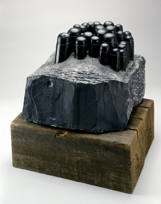 A black, stone-like structure with black cylindrical forms on top rests on a wooden platform.