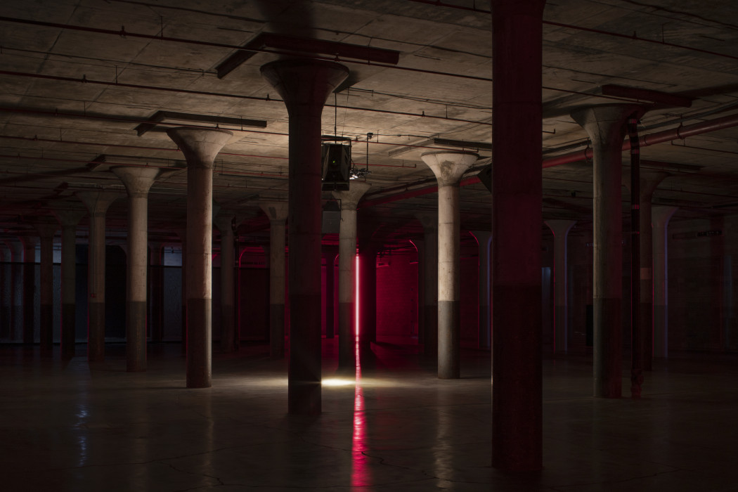 A dark industrial basement with cement columns is illuminated by a warm tone white spotlight focused on the floor. A bright red neon tube that peaks through the columns in the background.