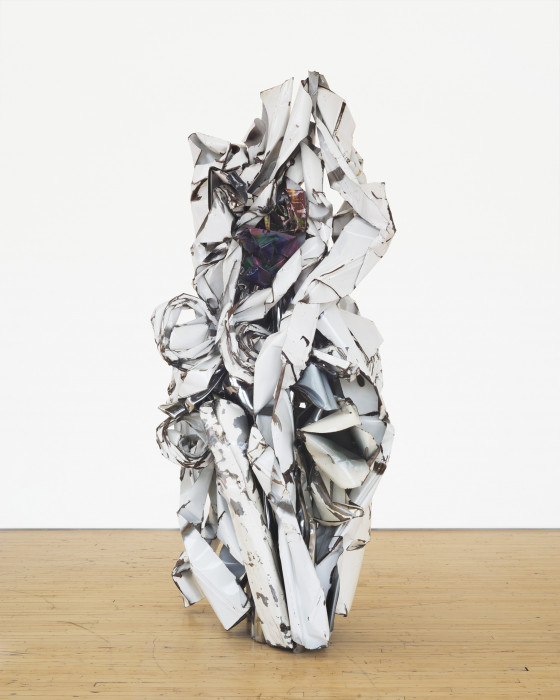 A vertically oriented sculpture made of white metal parts that are twisted around an iridescent metal center rests on a wooden floor.