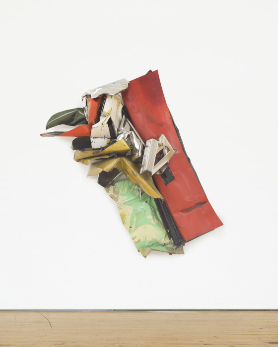 A diagonally mounted sculpture made red, yellow, green, and white metal parts hangs on a white wall.