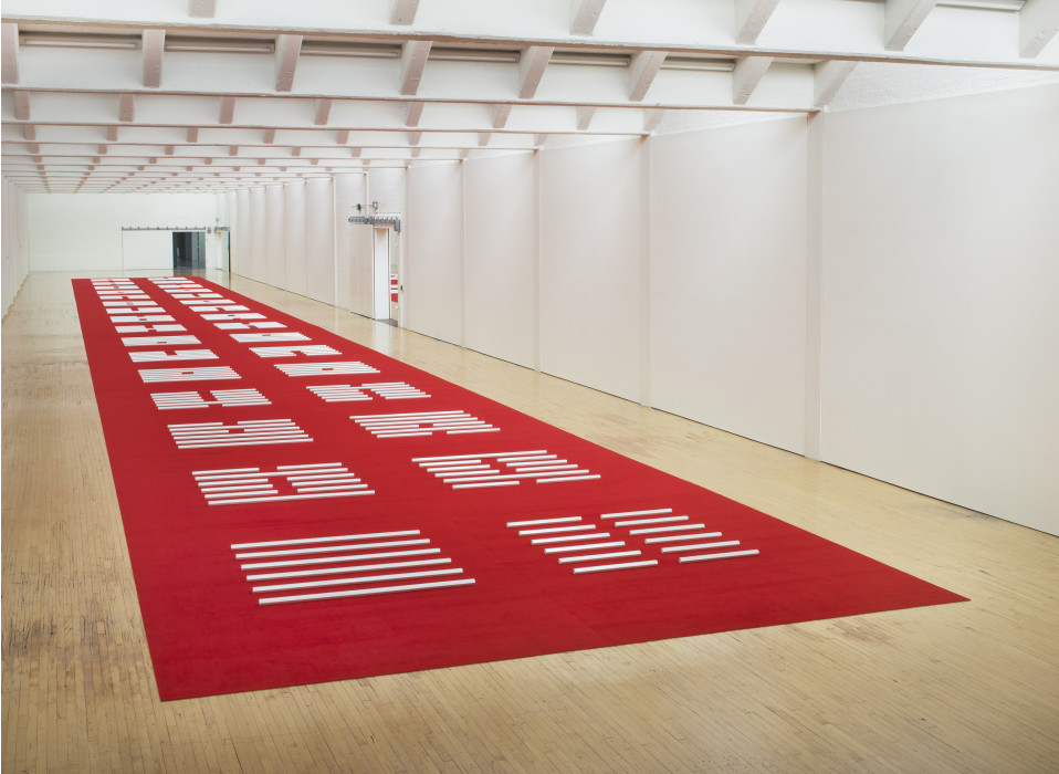 A series of many white hexagonal poles are horizontally placed on a red carpet that spans the length of a large white room with a wooden floor.