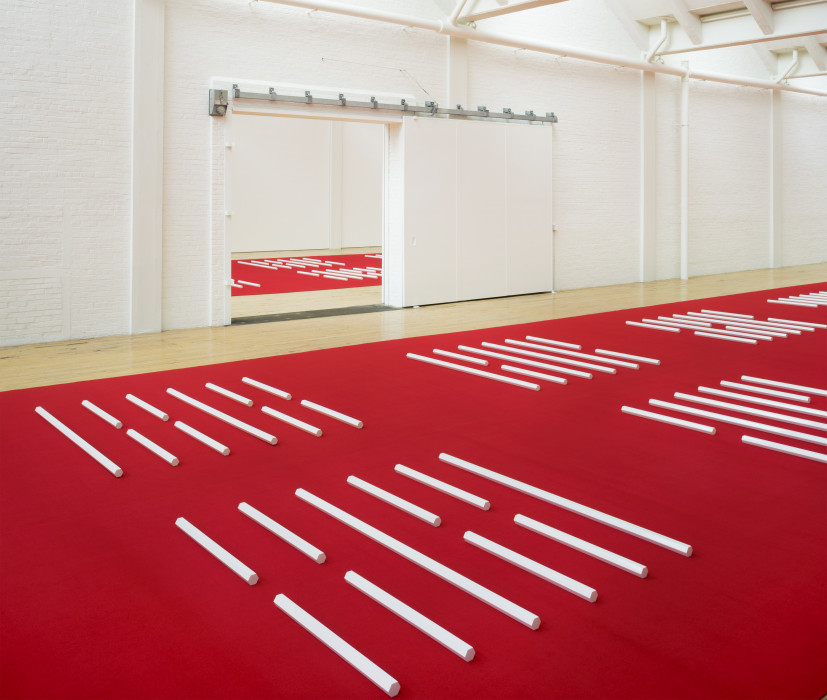 A series of white hexagonal poles are horizontally placed on a red carpet in a white room with a doorway that opens into another similar room.