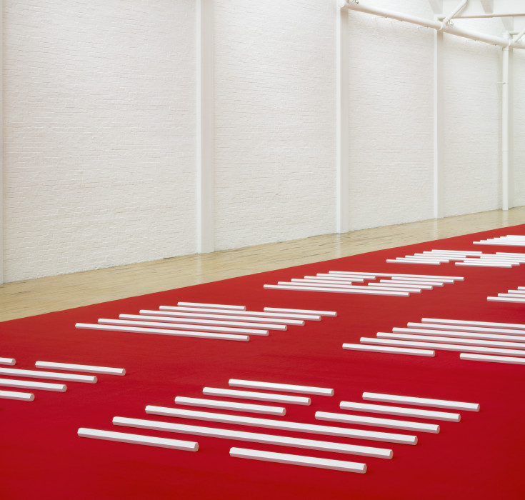 A series of many white hexagonal poles are horizontally placed on a red carpet that spans the length of a large white room with a wooden floor.
