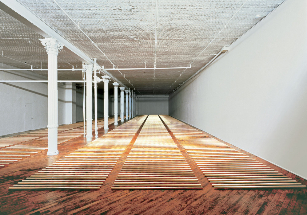 Five vertical columns of sections of brass rods on a wood floor.