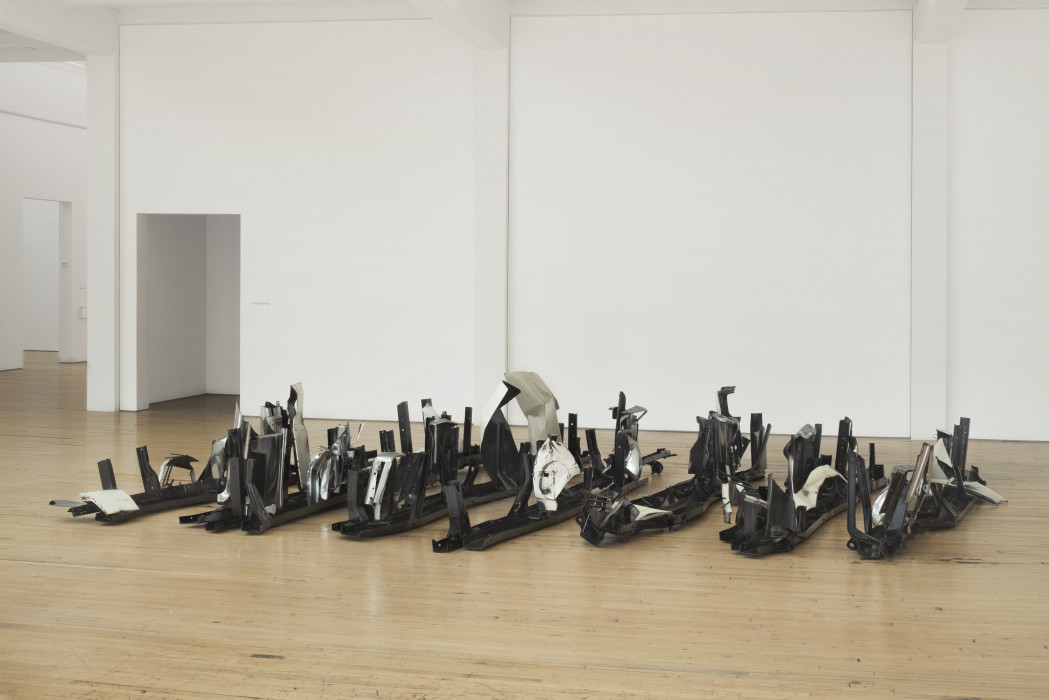A series of long, spiky, black, metal sculptures are arranged in parallel lines on a wooden floor.