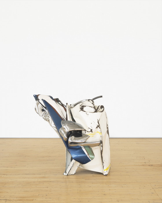 A squat sculpture made of chrome, white, and blue metal parts that twist together rests on a wooden floor.