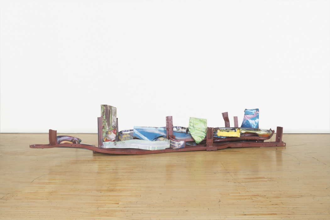 A long, low-lying sculpture made of red, blue, green, and multicolored metal parts rests on a wooden floor.