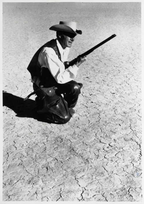 A cowboy holds a shotgun and kneels on a barren desert floor in this black-and-white film still.