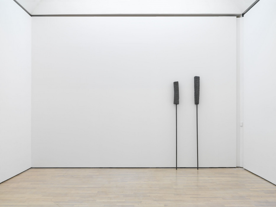 Two thin, grey rods with textured, different sized, irregular rectangular, grey forms on their top ends lean against a white wall in an empty gallery space.