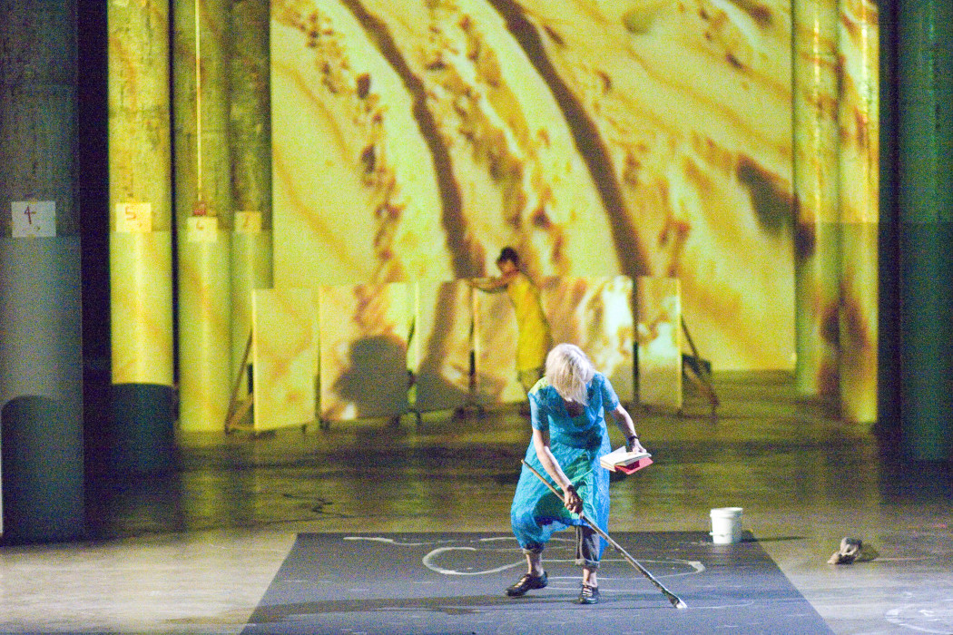 A woman wearing a blue dress stands in front of a yellow backdrop while looking at a book and painting curved white lines on the floor. 