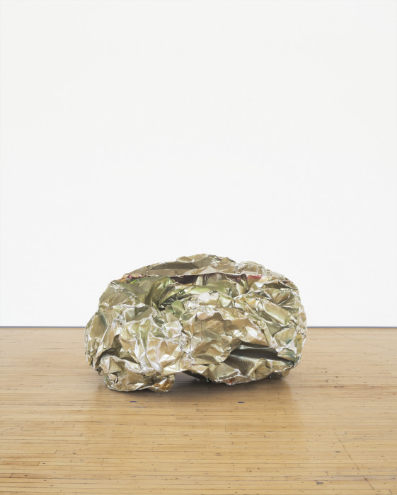 A crumpled ball-like sculpture made from reflective silvery green metal parts rests on a wooden floor.