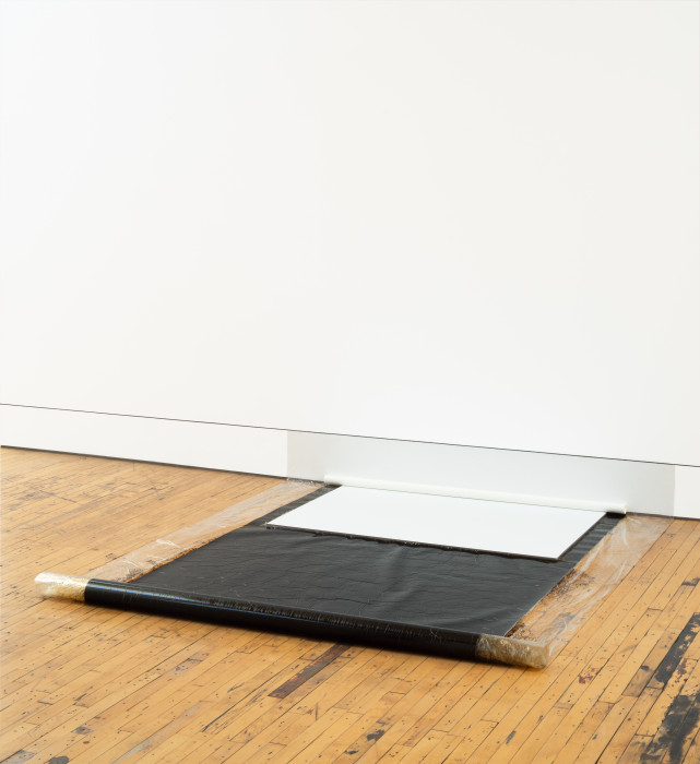 A roll of a rectangular-shaped, slightly transparent, and mostly black object extends from a white wall and rests on a wooden floor. A smaller, white, rectangular object also touches the wall and is placed above the black unrolled object.