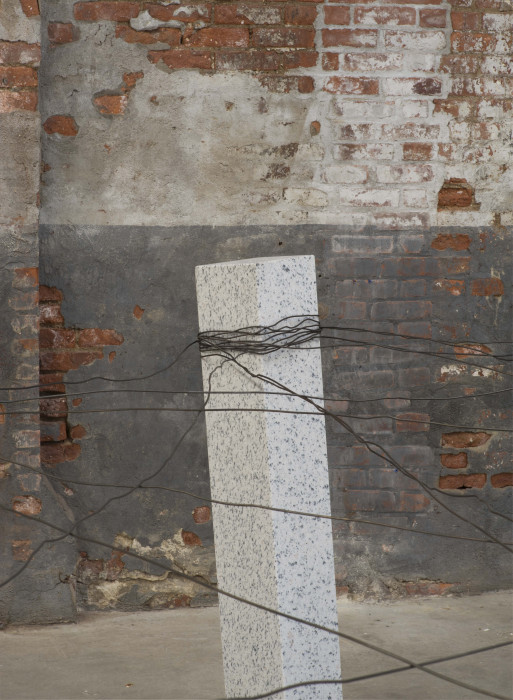An upright, rectangular block of speckled granite is wrapped with steel wire and stands in front of a brick wall.