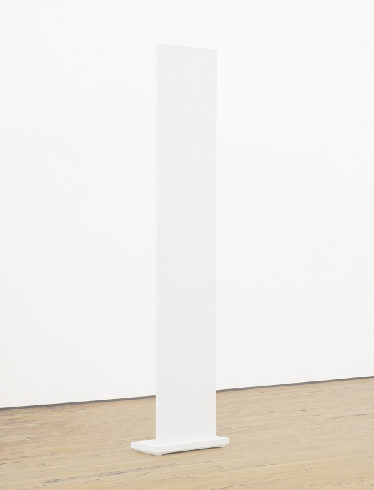 A vertically oriented, white panel with a small rectangular base is placed on a wooden floor in front of a white wall.