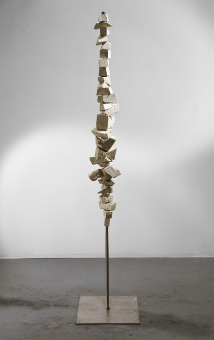 Balanced on a bronze pole and attached to a flat base is a thin vertical sculpture made of several wooden blocks of varying sizes that are stacked on top of one another.