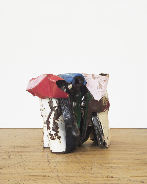 A squat sculpture made of red, pink, green, blue, and white metal parts rests on a wooden floor.