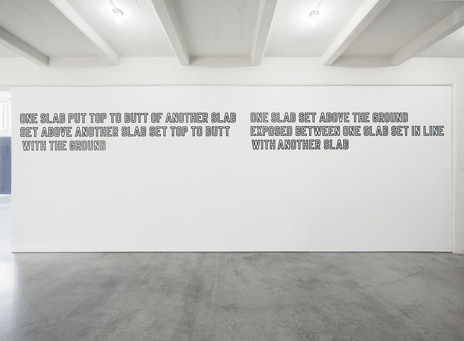 Two sets of white text outlined in black are placed high on a white wall. The text on the left reads: 