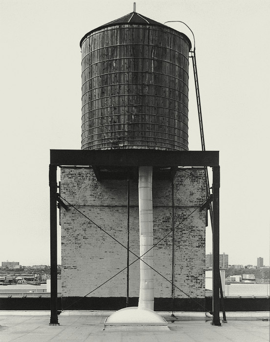 Black-and-white photograph of a rooftop water tower above a small brick penthouse surrounded by metal supports