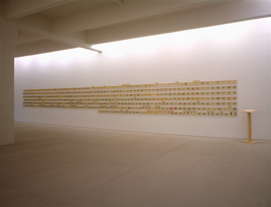Over four hundred framed, small color drawings hang in seven neat rows on a white wall with a thin wooden lectern sitting to the right of the drawings.