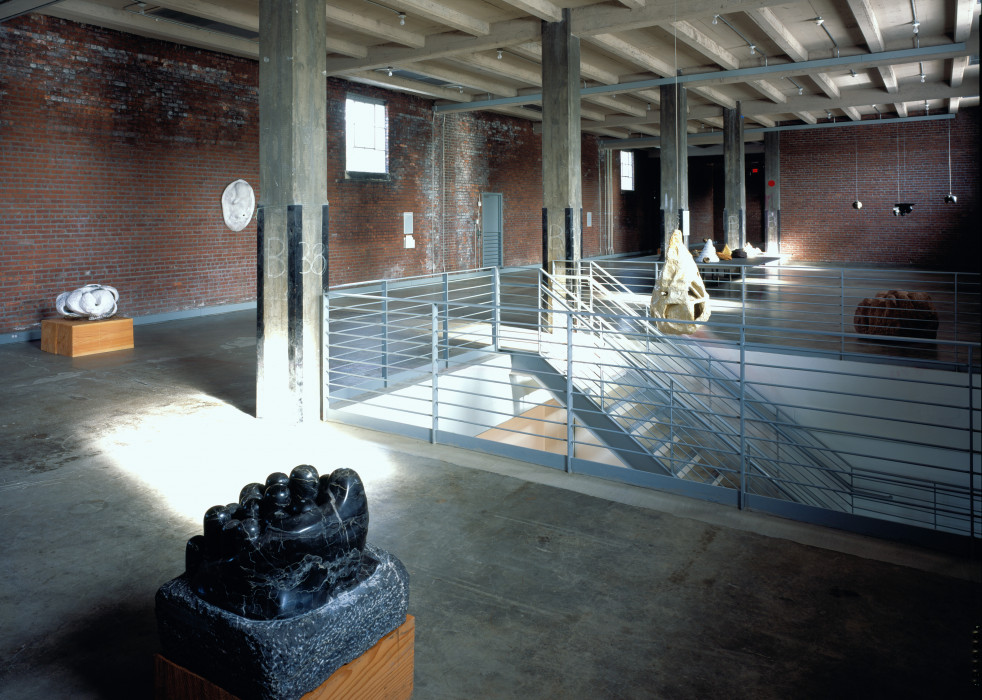 Many organically formed shapes rest on platforms, hang from wires, or protrude from the walls of this large, warehouse-like room with brick walls and cement ceiling, columns, and floor. A stairwell is placed within the center of the room and bright light streams through several windows.