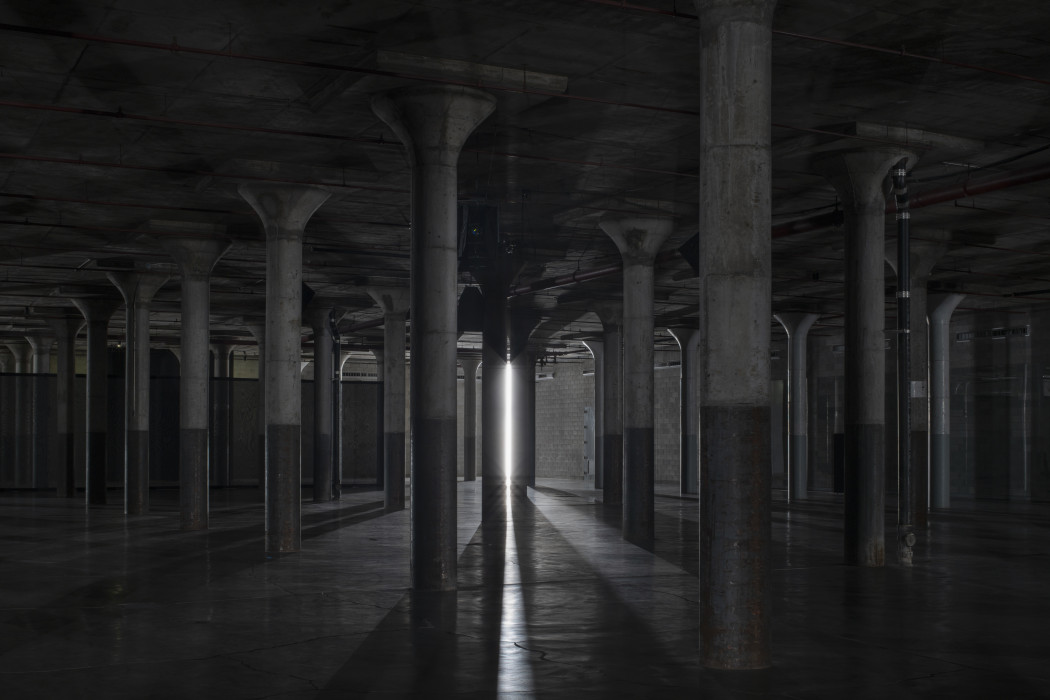 A dark industrial basement with cement columns is illuminated by a single bright vertical tube light that peaks through the columns in the background.