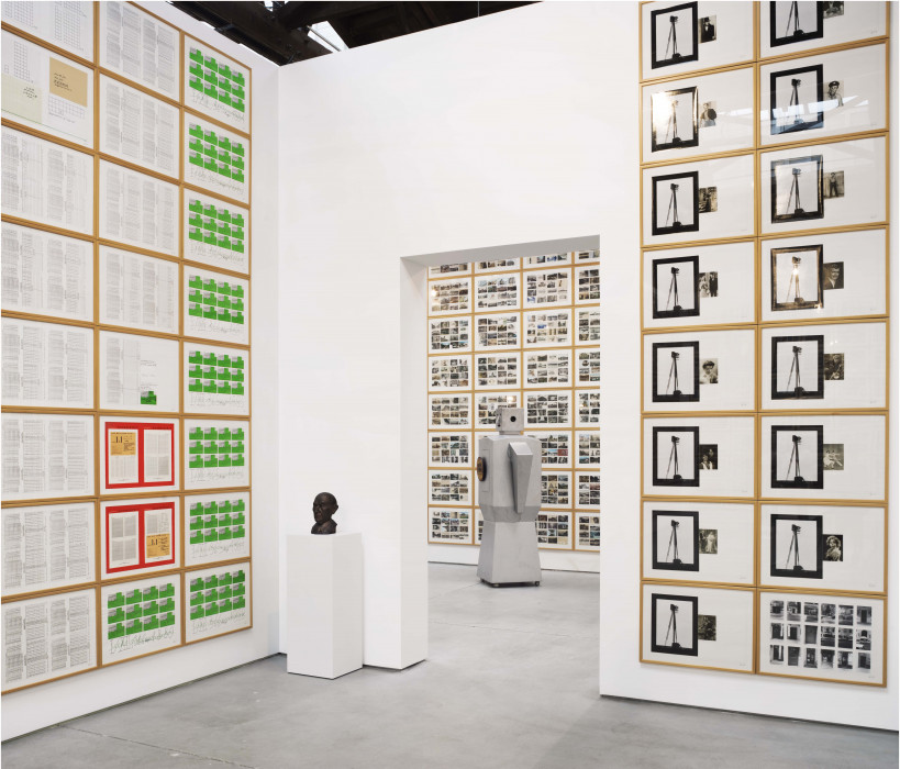 Paper works installed in a locked grid  on two walls, each with a white background, each with uniquely collaged images, texts, pages. postcards, or handwritten notes. An open doorway shows the work continuing into the room beyond. By the door is a pedestal with the sculpture of a man's head. In the distant room, a robot sculpture stands on the cement floor.