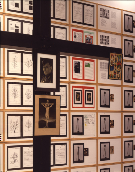 Paper works installed in a locked grid, each with uniquely collaged images, texts, pages. postcards, or handwritten notes.  A black cross in front of the wall has 3 framed photographs attached, a photo of a man in military attired, a drawing of Jesus on a cross, and an illustration of children's dolls.