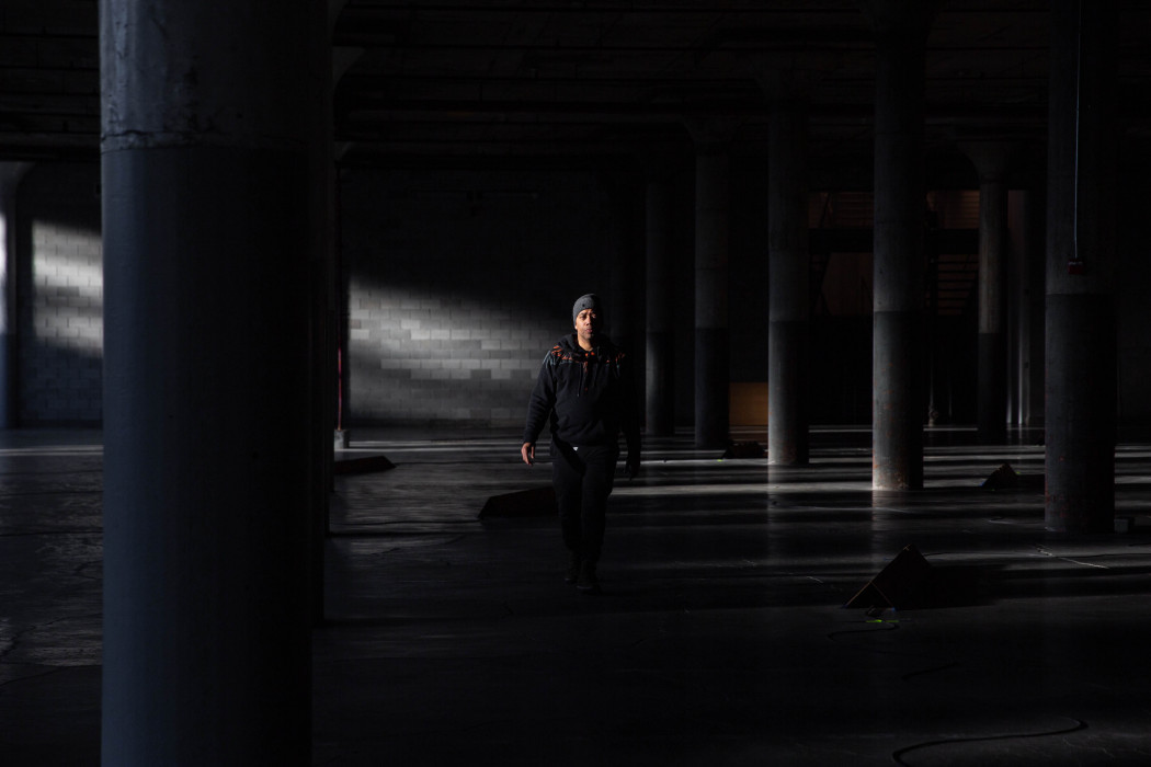 A person in black clothes and a grey beanie walks forwards in a dark industrial basement and is illuminated by natural light.