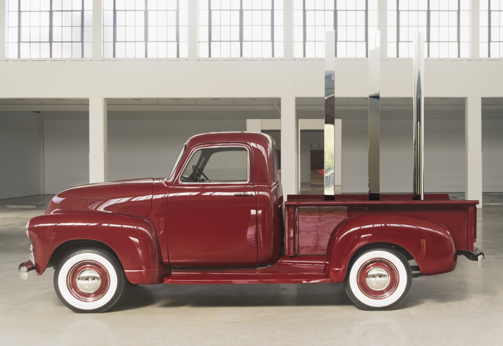 A vintage red Chevrolet pickup truck sits in profile in an open gallery space with a row of large windows in the background. Placed vertically in the truck's flatbed are circular, square, and triangular polished-stainless-steel rods.