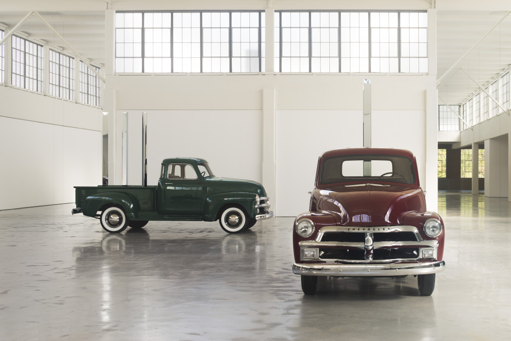 Two vintage Chevrolet pickup trucks, one red, one black,  in an open gallery space with a row of large windows in the background.  Placed vertically in the trucks' flatbeds are circular, square, and triangular polished-stainless-steel rods.