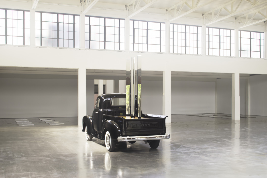 The back view of vintage black Chevrolet pickup truck in an open gallery space with a row of large windows in the backgroundront of a white wall. Placed vertically in the truck's flatbed are circular, square, and triangular polished-stainless-steel rods.