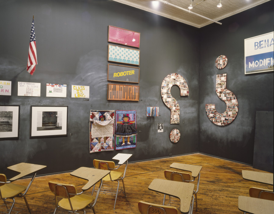 A classroom with wooden floors and black chalkboard walls, containing seven wooden chairs with attached desks, facing toward two large question marks hung on either side of a corner of the room, one upside down. Also hung on the walls are various drawings, small quilts, framed posters, photographs, a book, and an American flag on a flagpole.