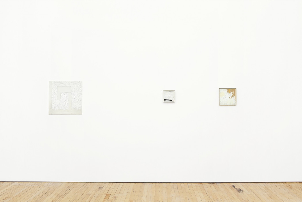 Three small, square white paintings hang on a white wall above a wood floor.