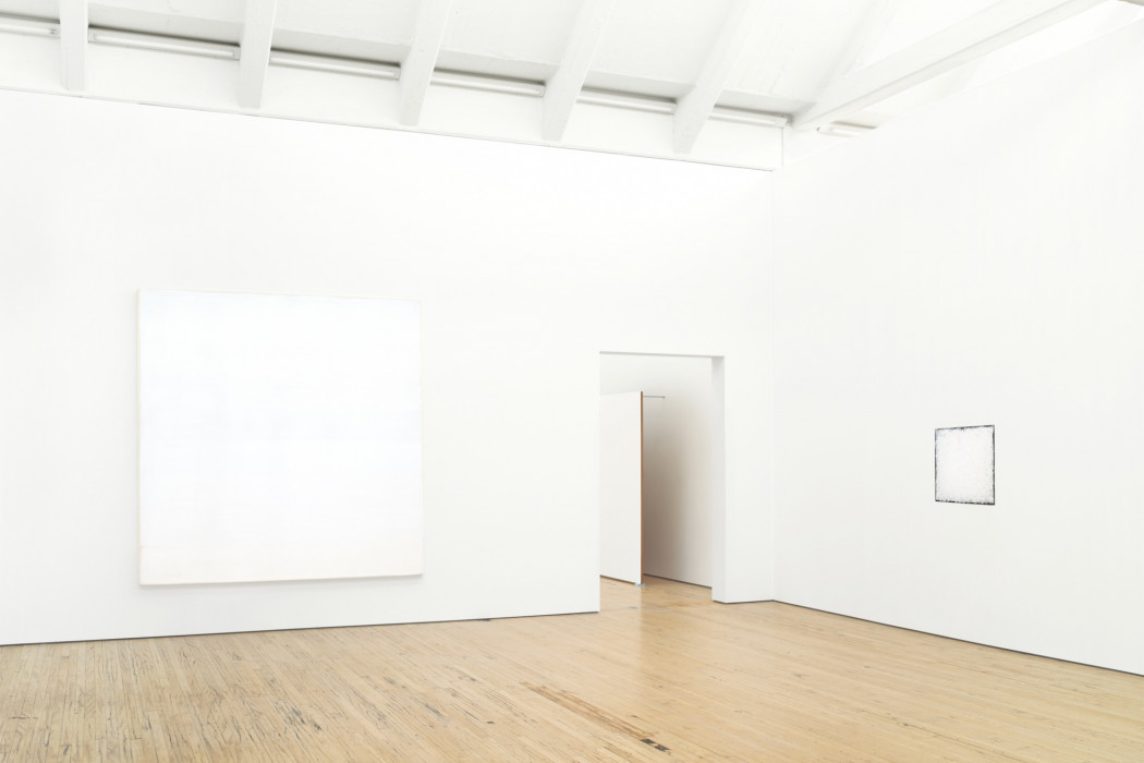 Two square white paintings hang on white walls above a wood floor. The one on the left wall and the right one is small. A large, rectangular white painting affixed to a wall by long metal rods and resting on the wood floor is partially visible through a doorway between the paintings.
