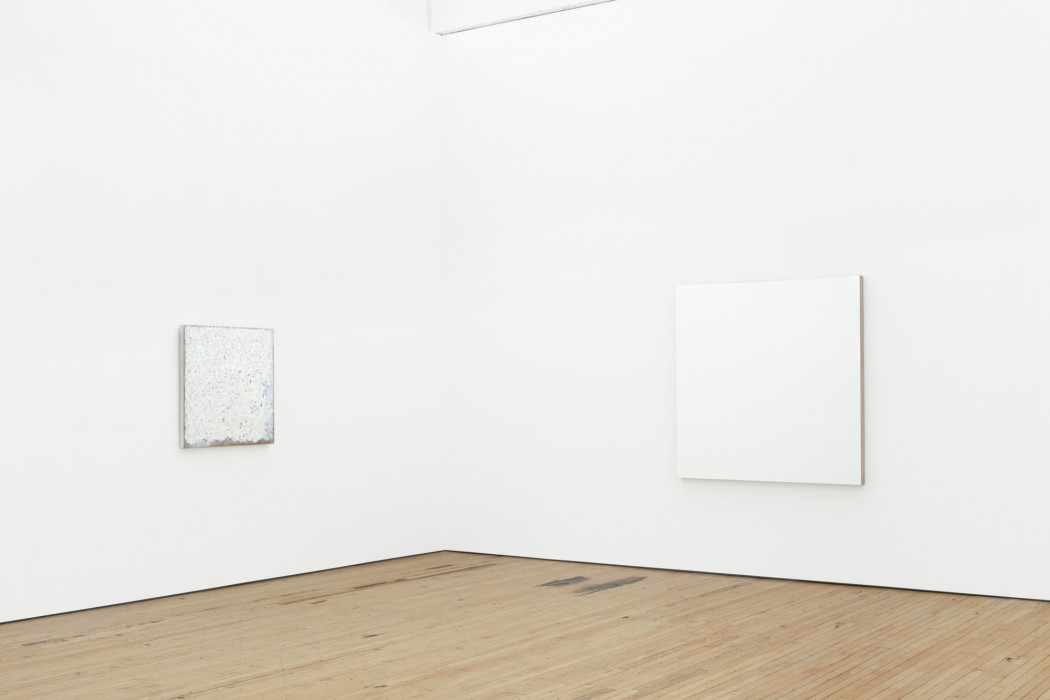 Two square white painting hang on walls above a wood floor on either side of a corner. The right one is large, and the left one is small.