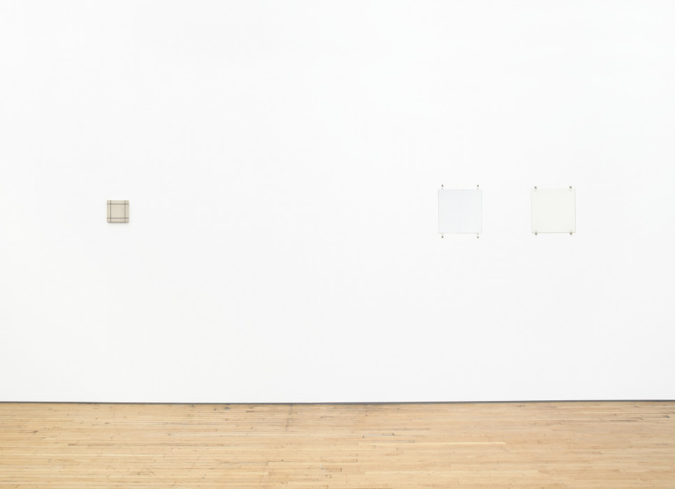 Three square paintings hang on a white wall above a wood floor. The far left painting is small and tan with a grid of black lines. The two white paintings on the right hang close together and are affixed with fasteners and bolts.